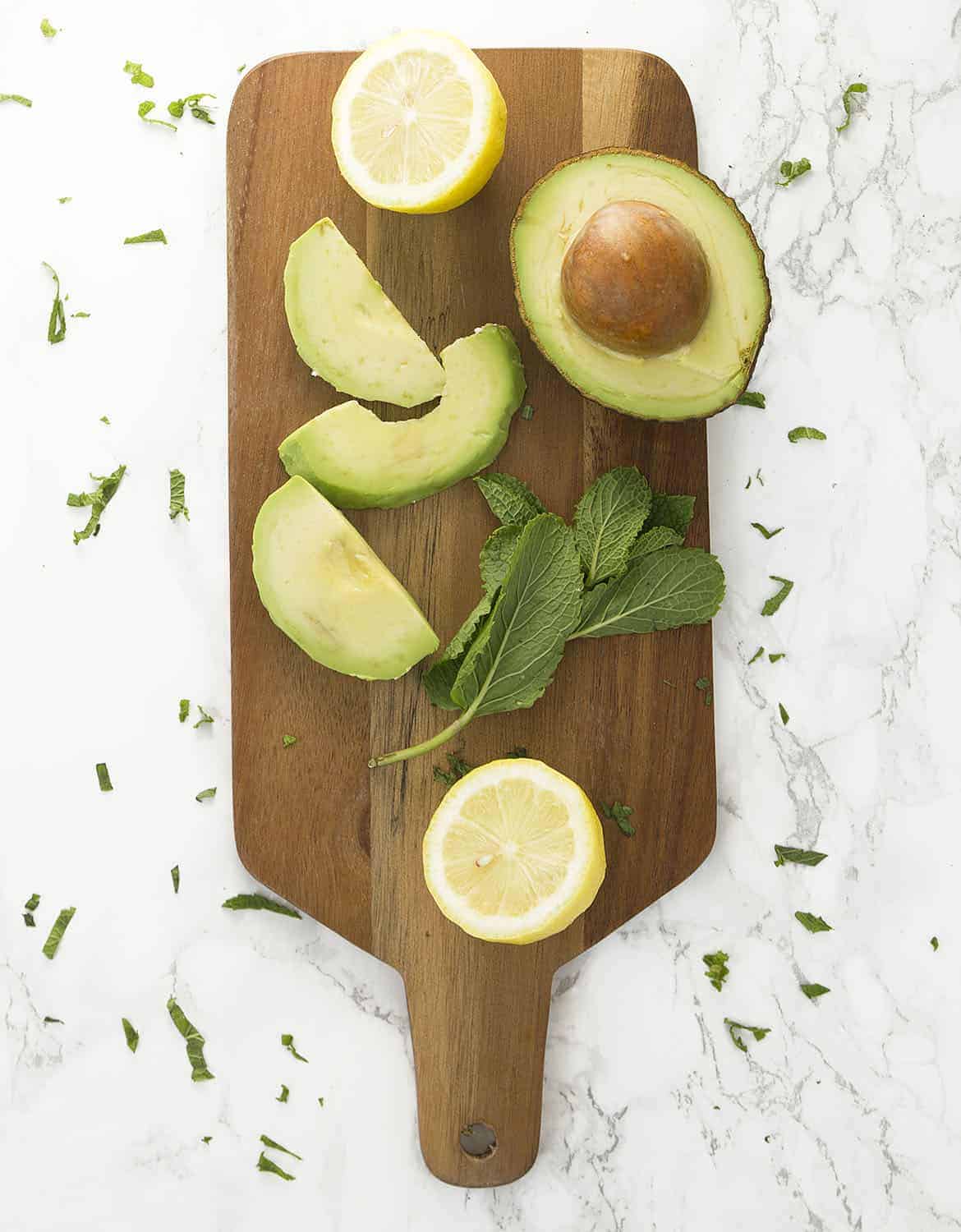 Half avocado, two lemon halves and mint leaves on a wooden chopping board.