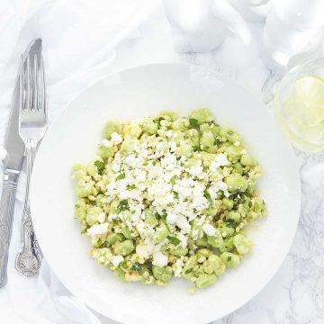 A green fava bean and barley salad with crumbled feta on a white plate.