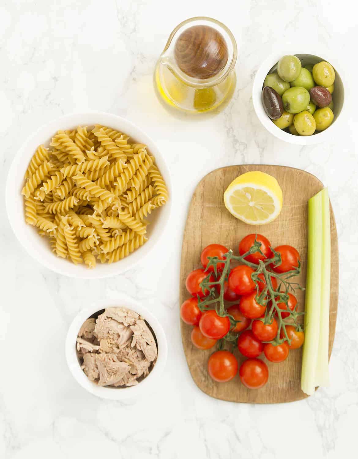 The ingredients for this tuna pasta salad are arranged on a white marble table and a wooden chopping board.