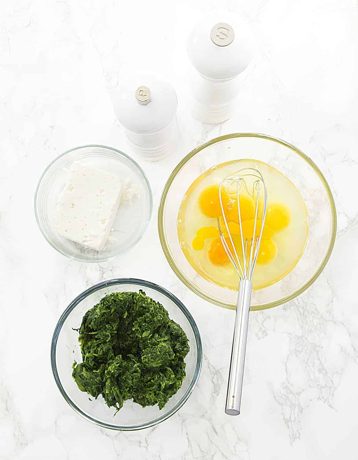 Three glass bowls with eggs, spinach and feta on a white marble background. 