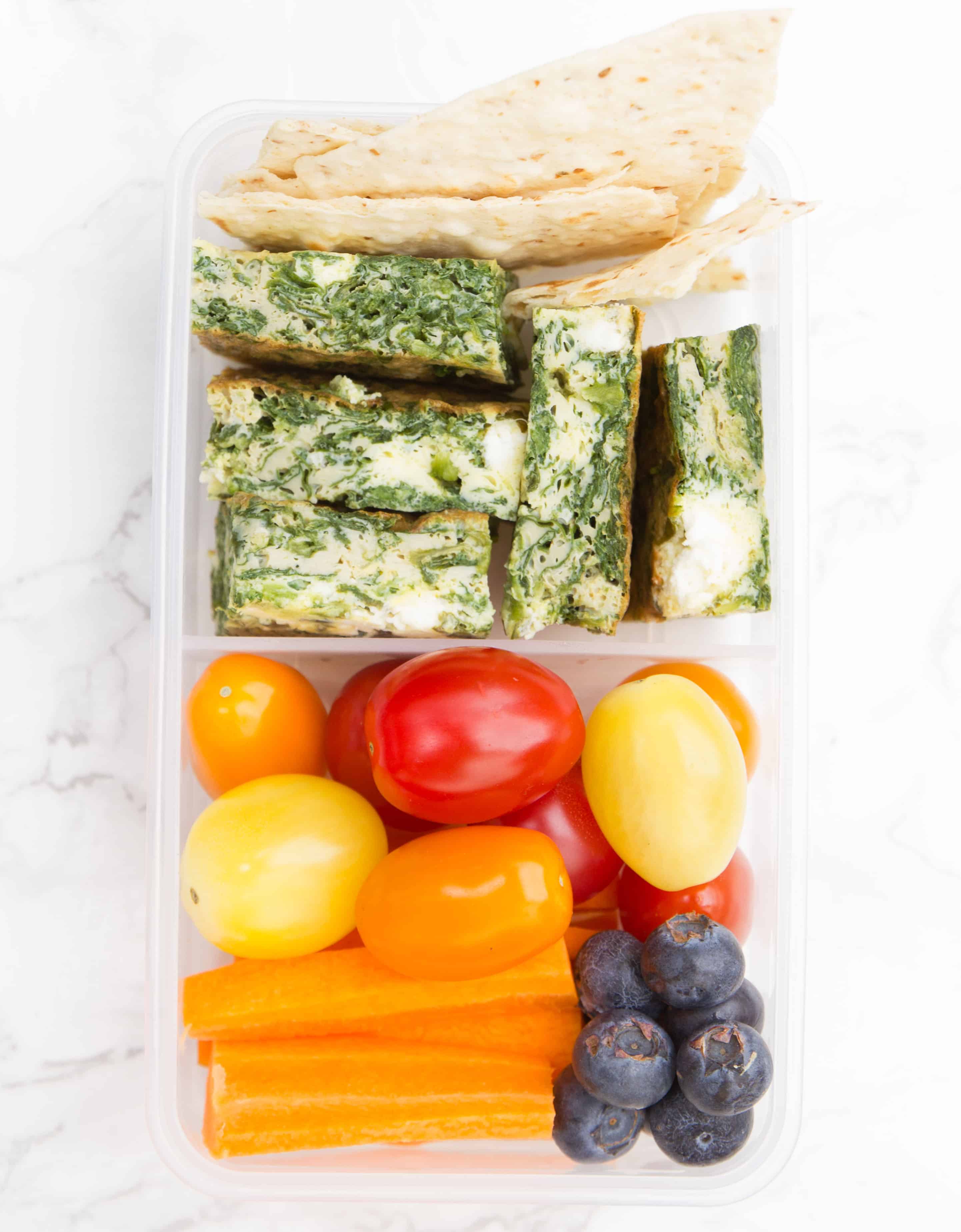 Cubes of spinach frittata in a rectangular meal prep container with vegetables and blueberries.