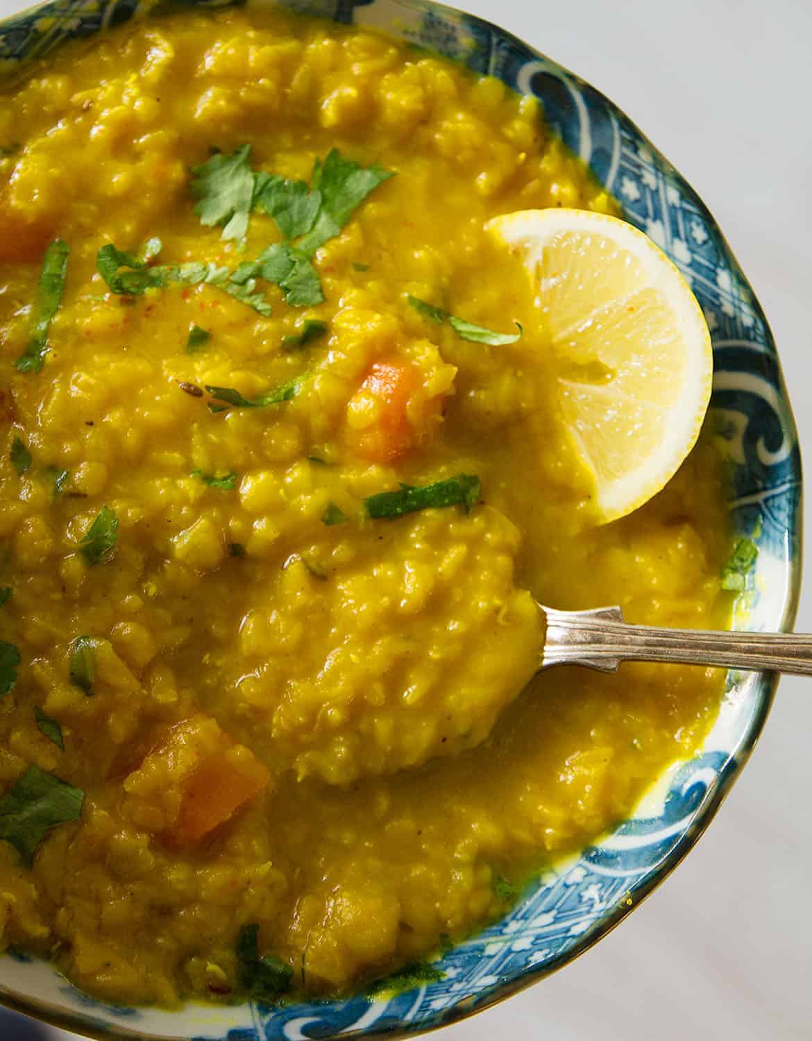 Red lentil soup in a blue bowl with a lemon wedge and a spoon over a white background.
