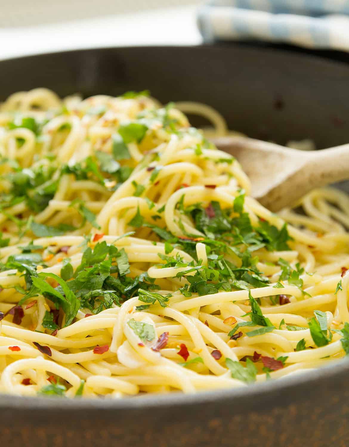 A wooden spoon is stirring spaghetti aglio e olio in a cast iron skillet.