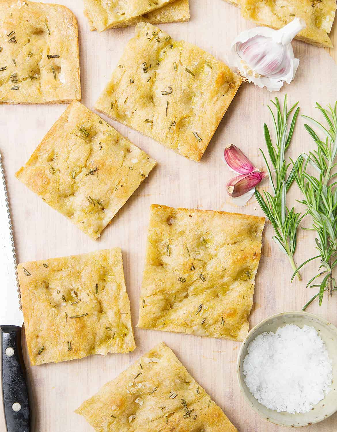 Slices of vegan focaccia with garlic cloves and rosemary on a chopping board - The Clever Meal