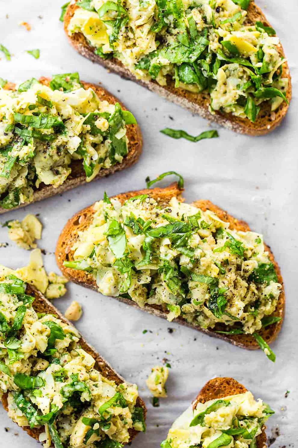 Five white bean artichoke basil toasts on white parchment paper - Pinch of Yum