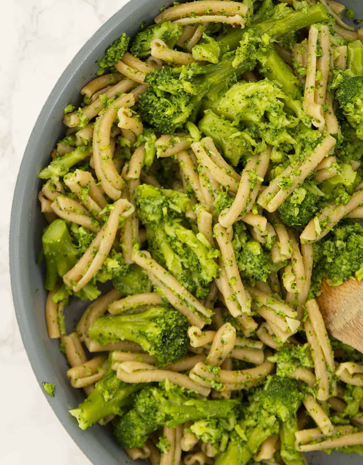 The broccoli pasta is stirred in a grey pan with a wooden spoon.