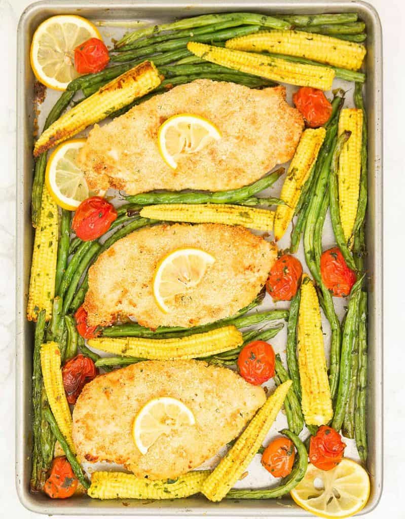 Top view of garlic chicken with lemon and roasted veggies over a baking tray.