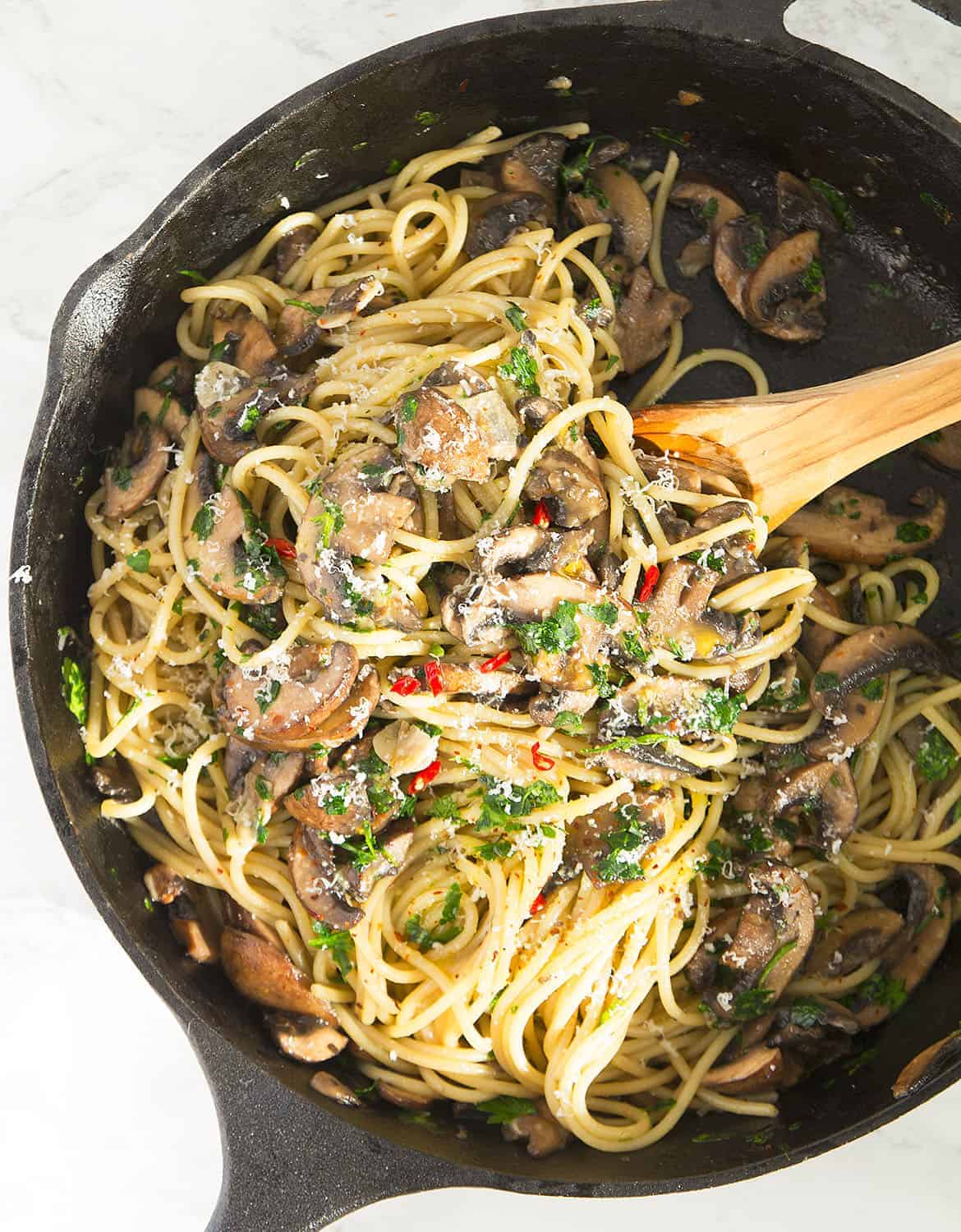 A wooden spoon is stirring the garlic mushroom pasta in a cast iron skillet.