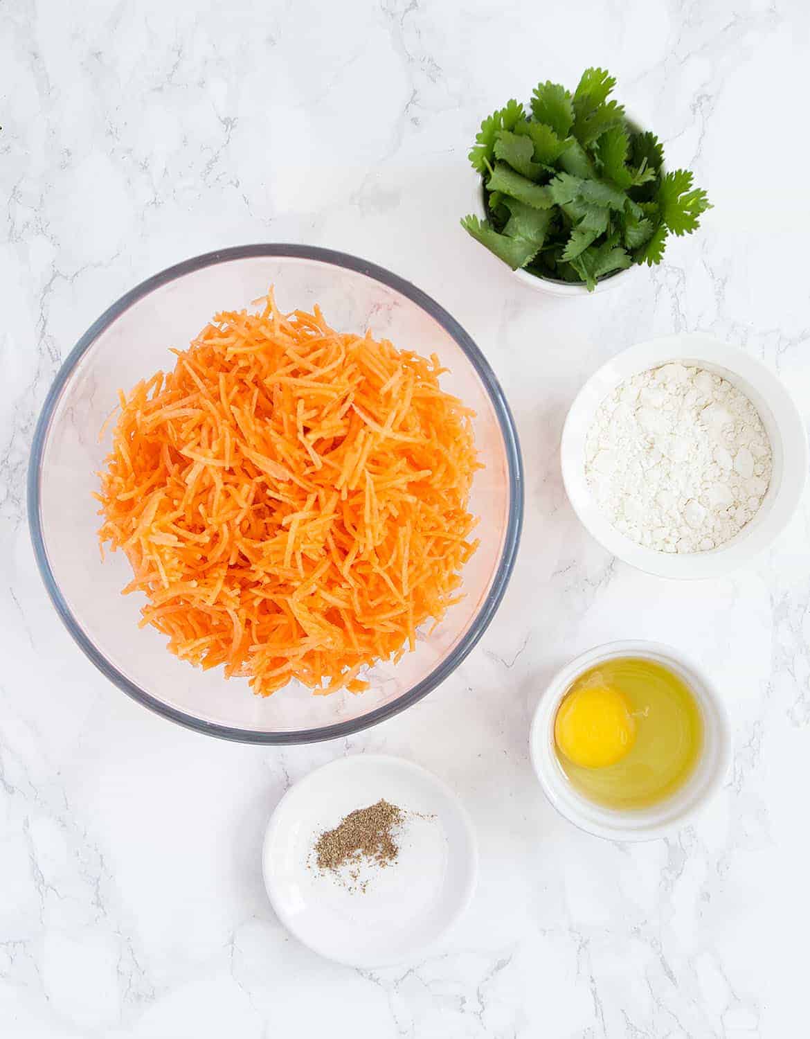 The ingredients to make carrot fritters are arranged on a white marble table.