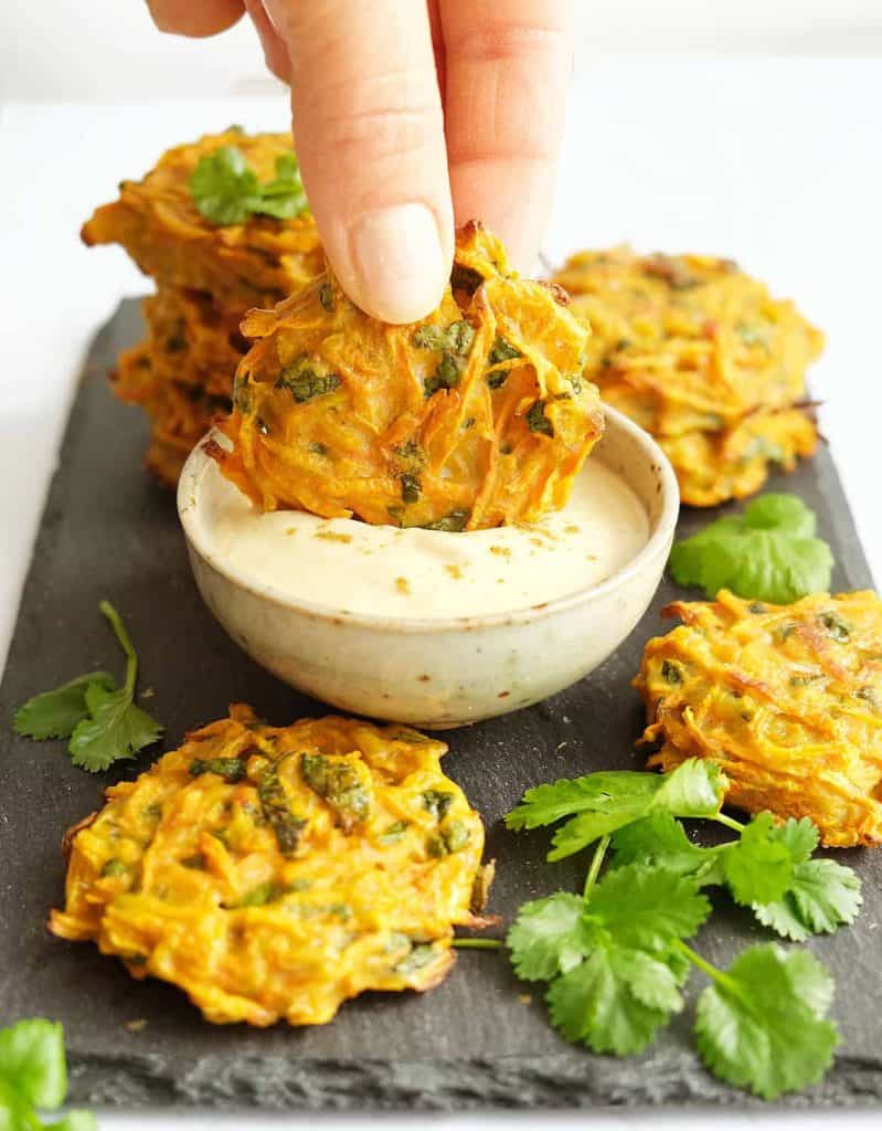 A hand dipping one of some carrot fritters into hummus dip.