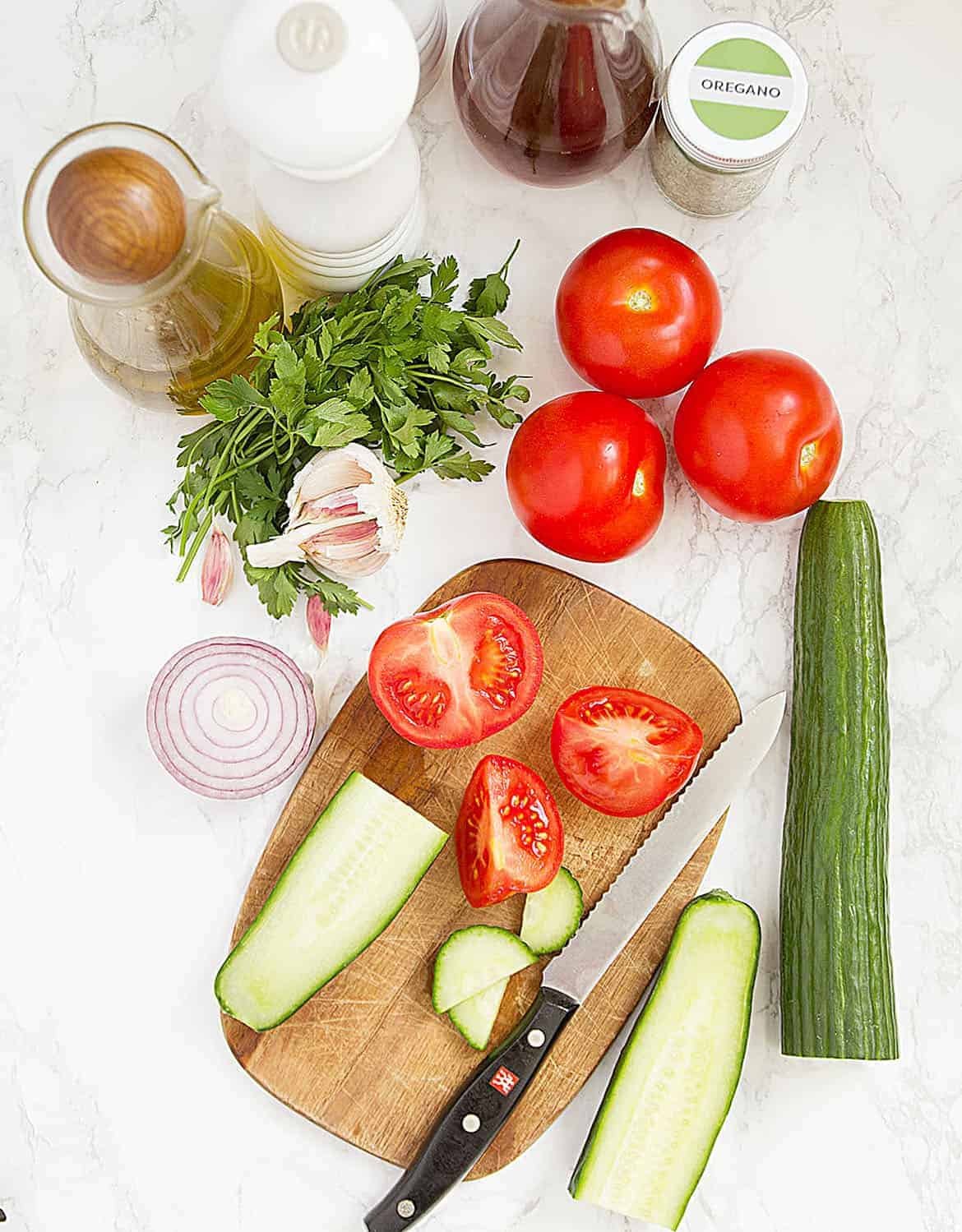 The ingredients for this tomato and cucumber salad are arranged around a chopping board over a white background.