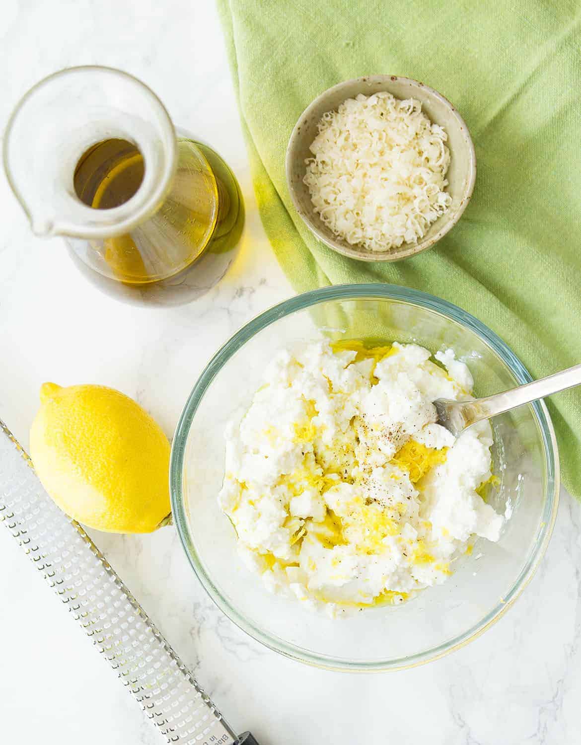 Ricotta, lemon zest, olive oil and parmesan are mixed with a spoon on a glass bowl.