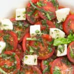 Close-up of marinated tomatoes, cubed mozzarella and fresh herbs on a white plate.