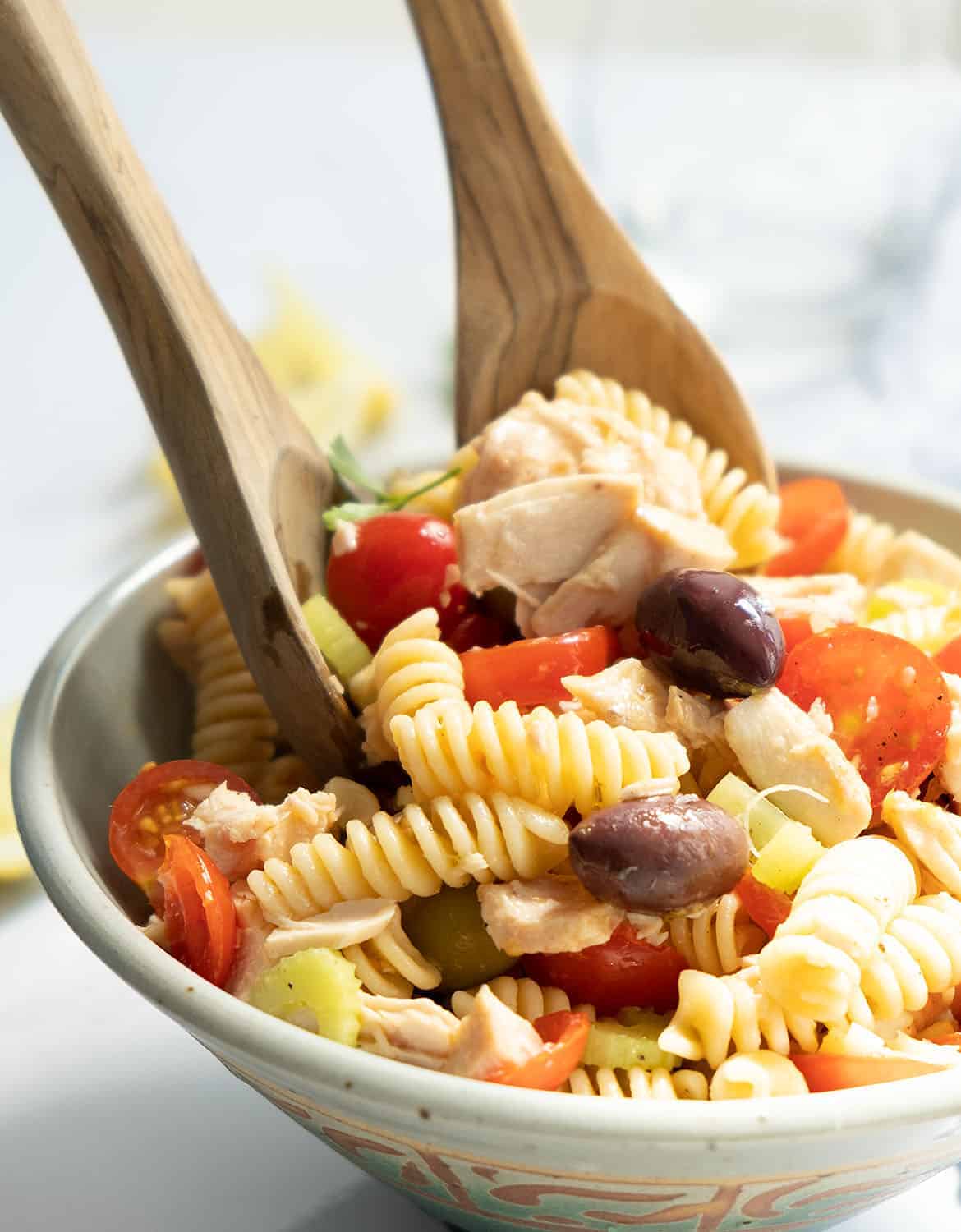 Two wooden spoons are mixing the tuna pasta salad in a bowl.