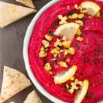A bowl full of beetroot hummus decorated with slices of lemon and chopped walnuts over a black background.