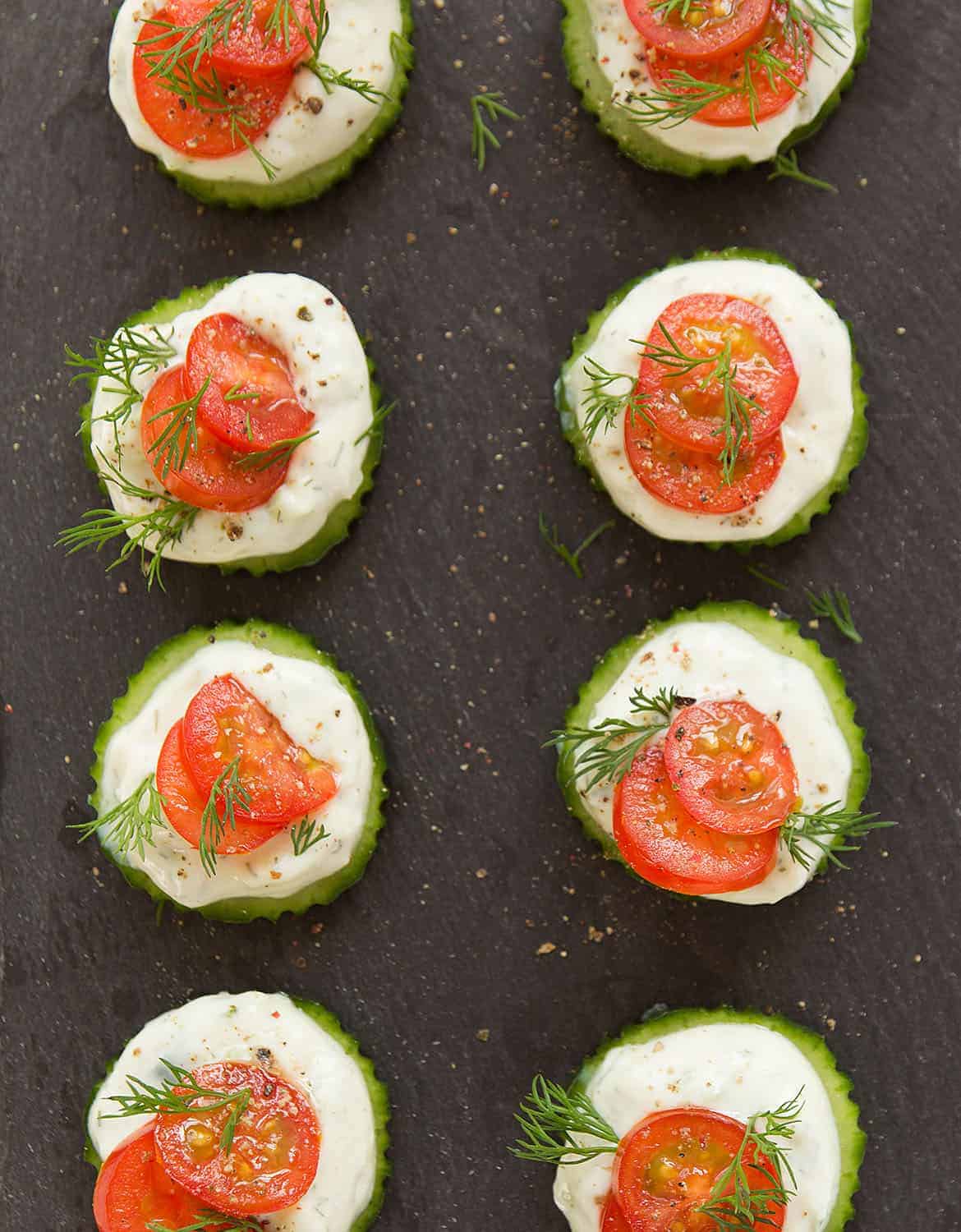 Eight slices of cucumber topped with vegan tzaziki and cherry tomatoes on a black tray - The Clever Meal