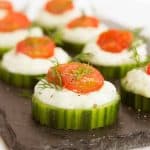 Slices of cucumber topped with vegan tzatziki and cherry tomatoes served on a black tray.