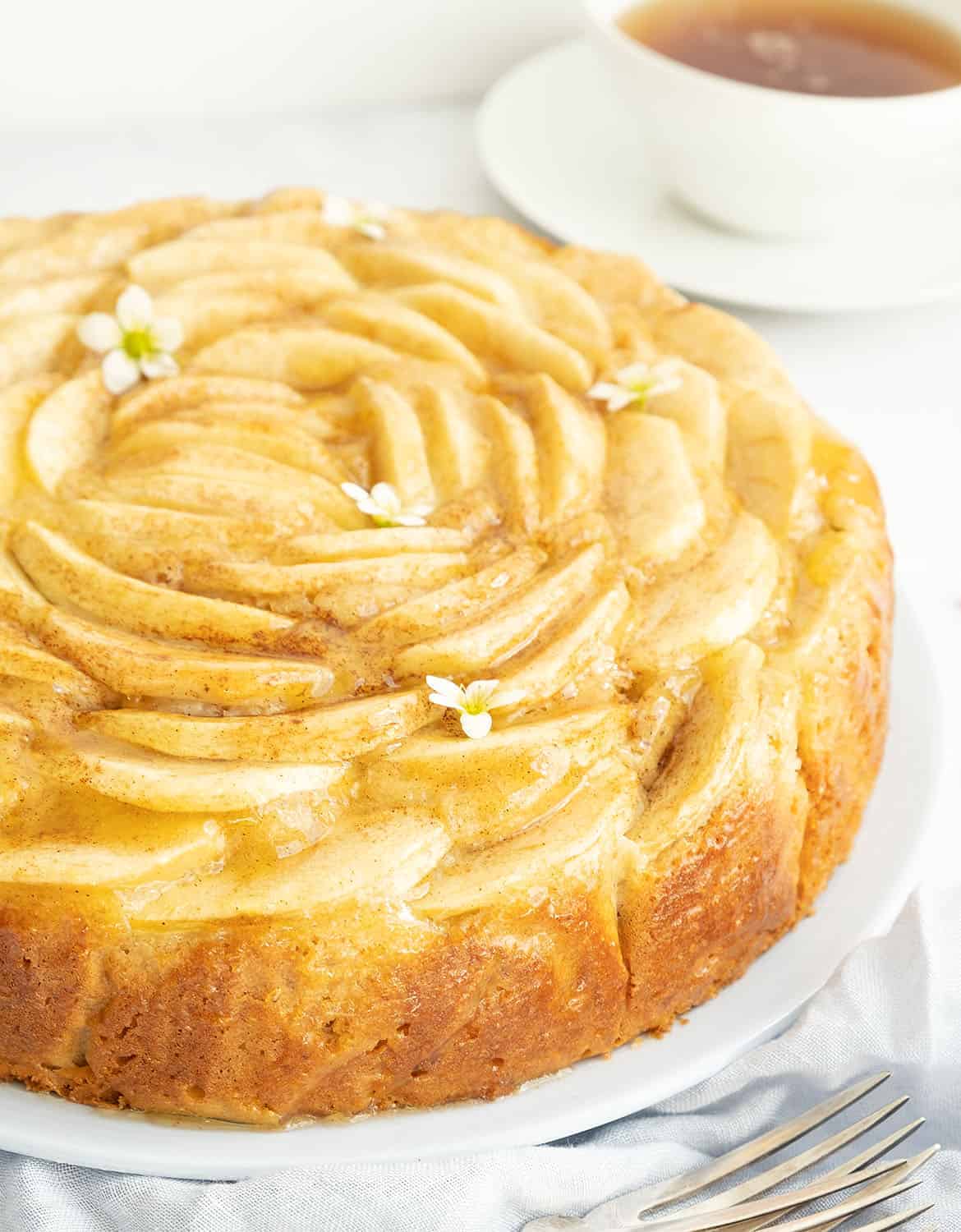 A close-up of a round apple cake decorated with small white flowers, a cup of tea in the background.
