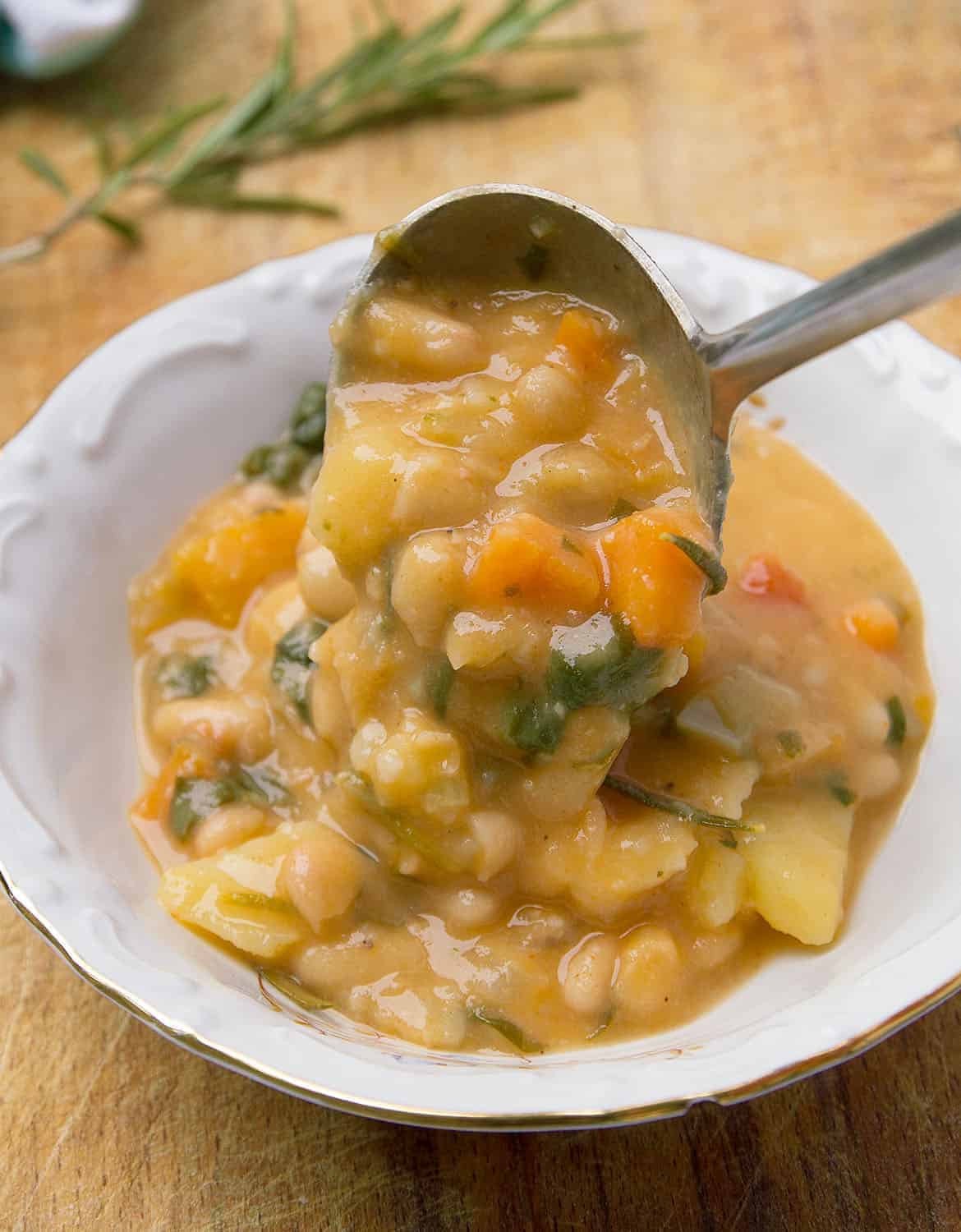 A ladle pouring some thick white bean soup into a white bowl, a sprig of rosemary in the background.