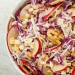 A white bowl full of cabbage salad with apple slices and walnuts.
