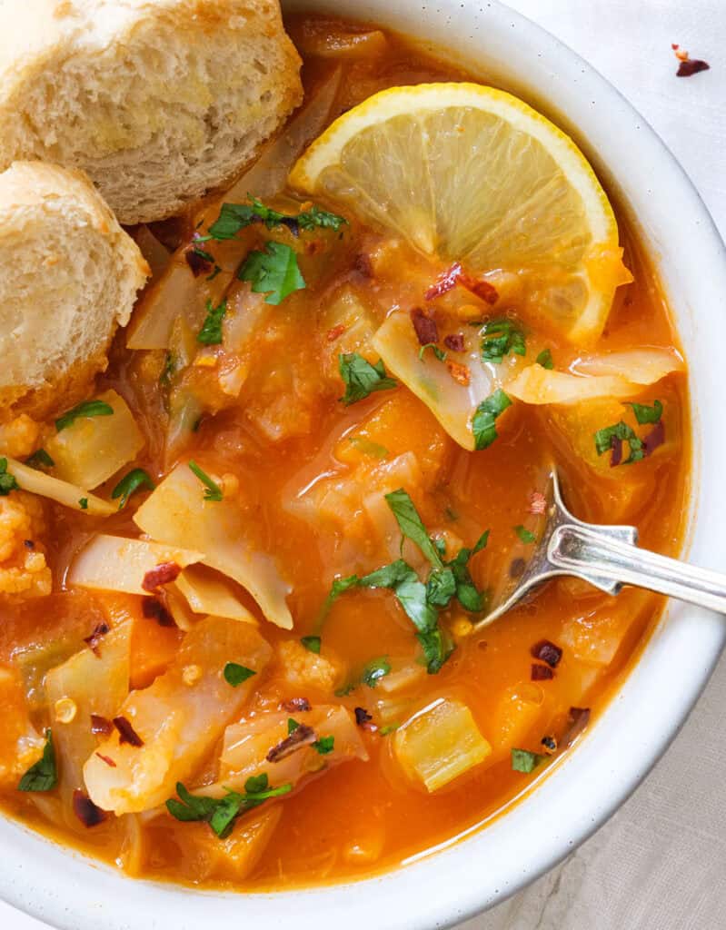 Close-up of a white bowl full of vegetarian cabbage soup served with bread and a lemon wedge.
