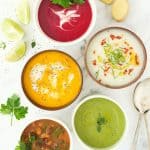 Five bowls of colorful weight loss soups over a white marble background.