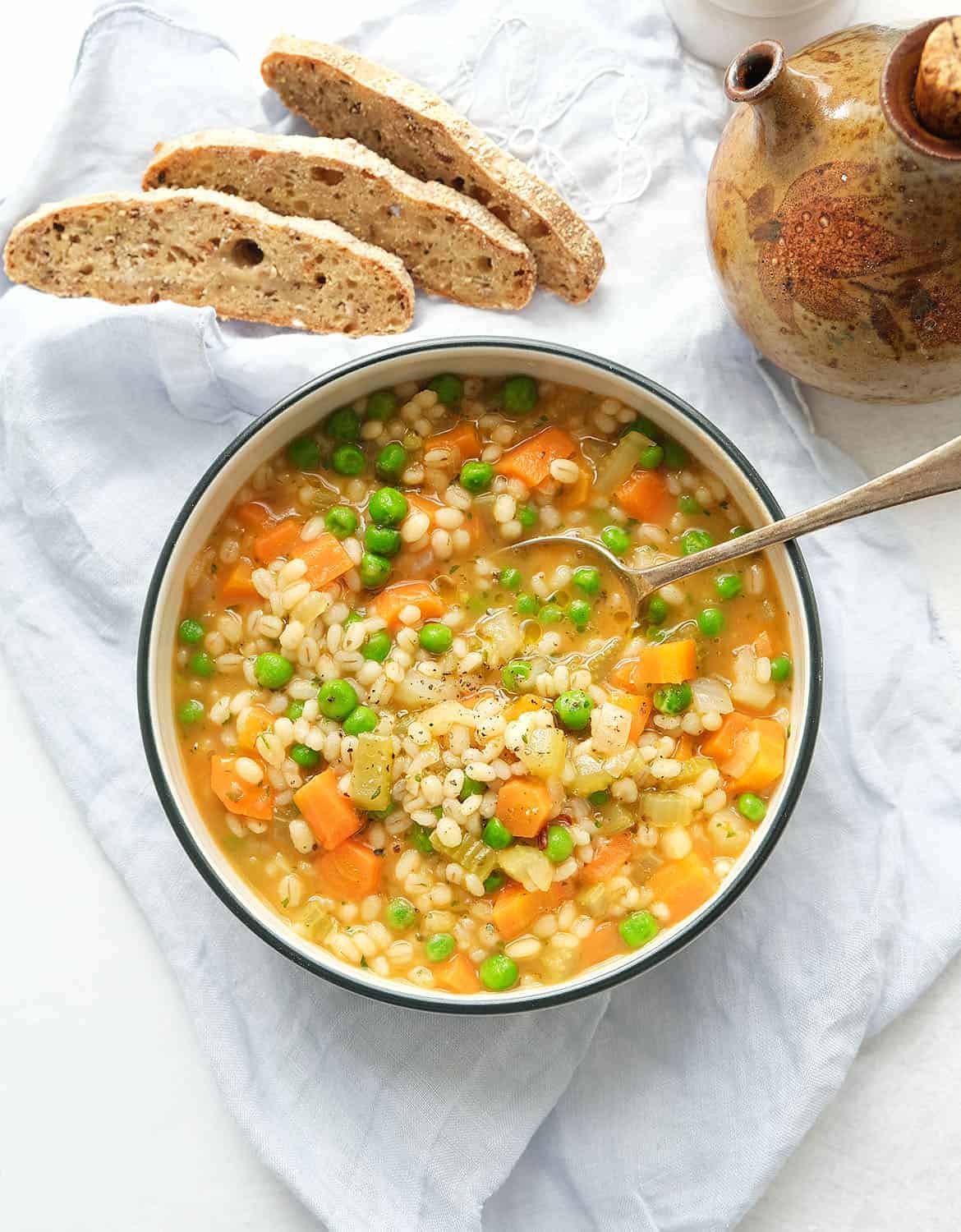 Vegetable barley soup in a white bowl with a spoon. Three slices of bread next to it.