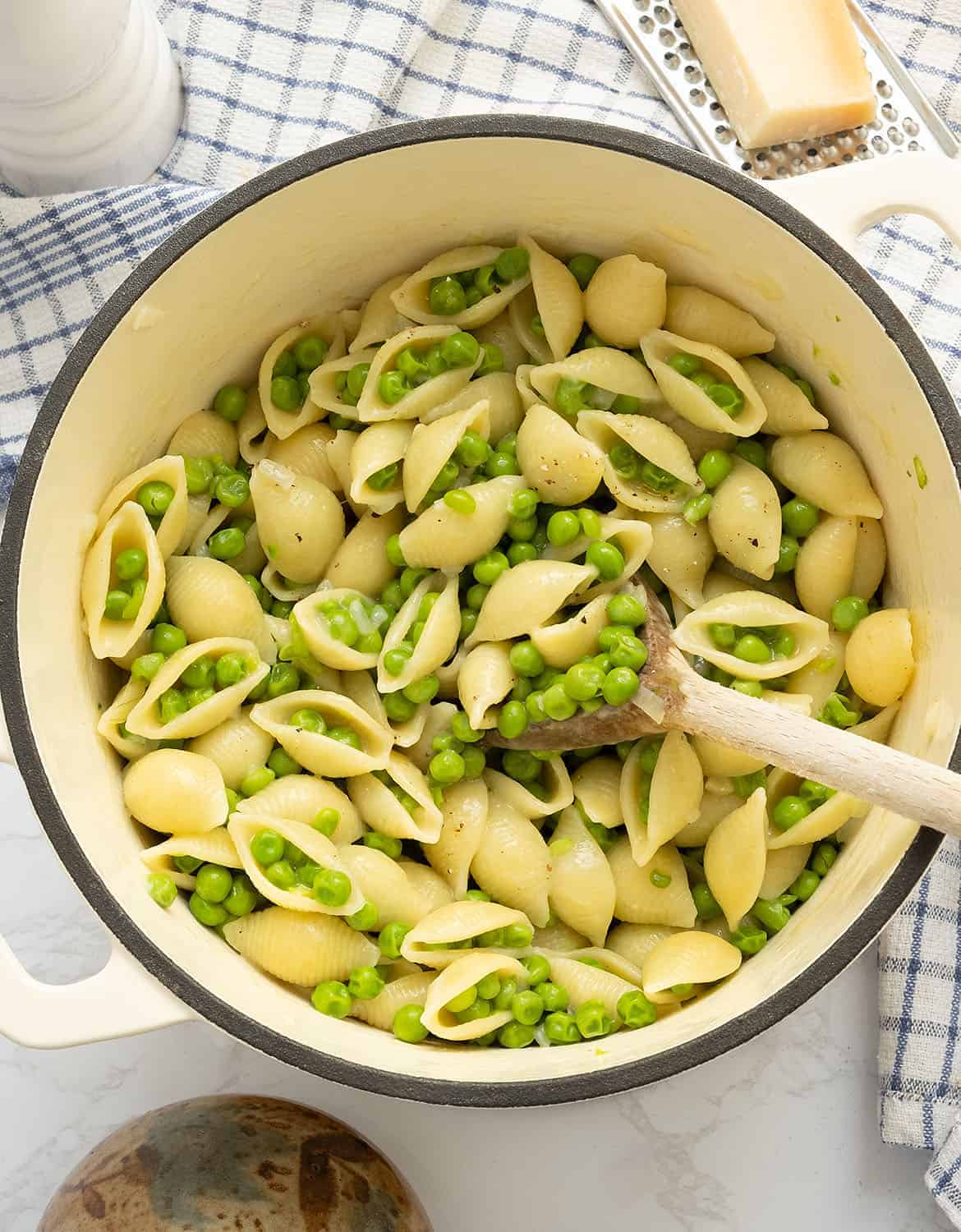 A wooden spoon stirs pasta with peas in a white cast iron pot.