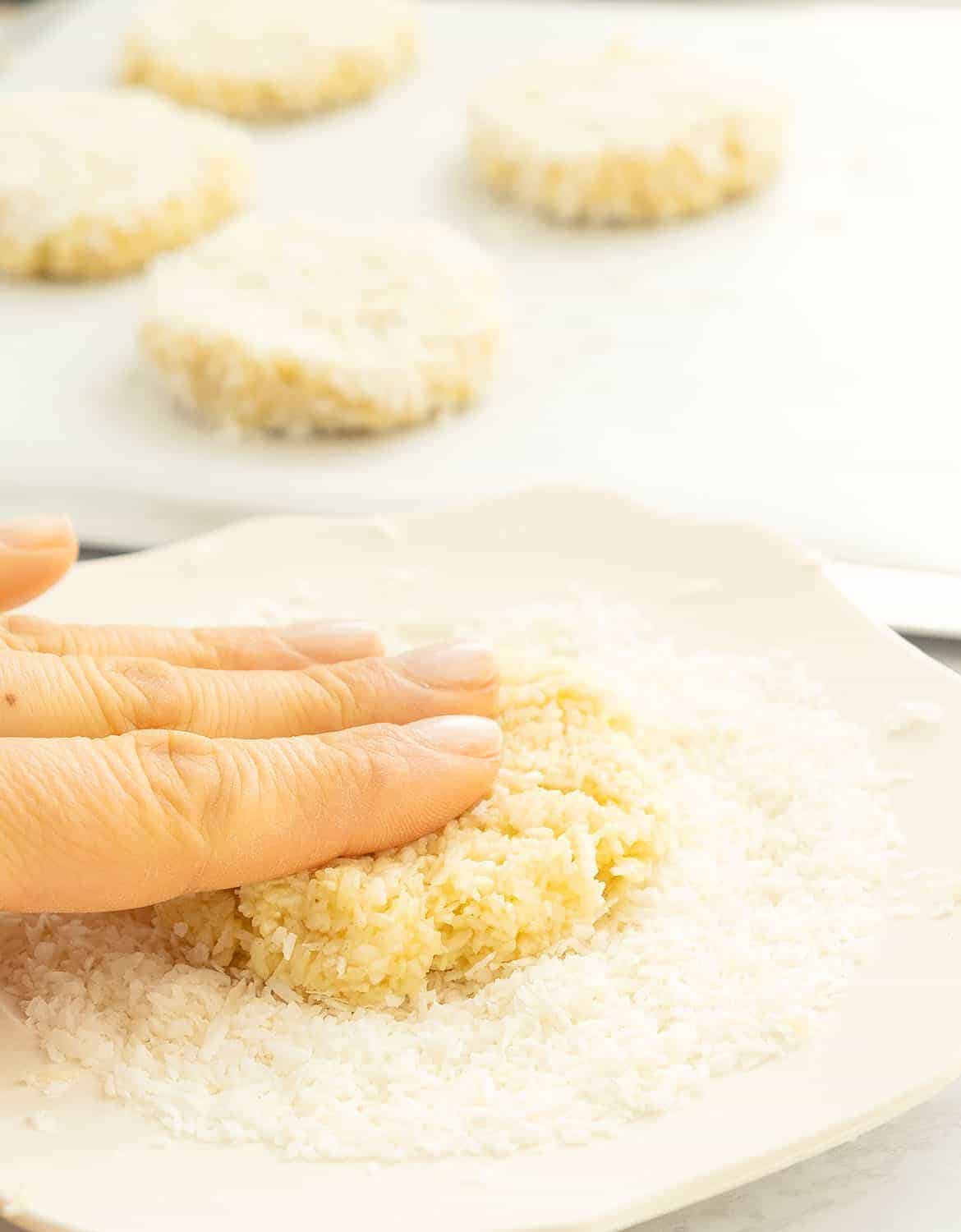 A hand pressing one coconut cookie on a plate full of shredded coconut. 