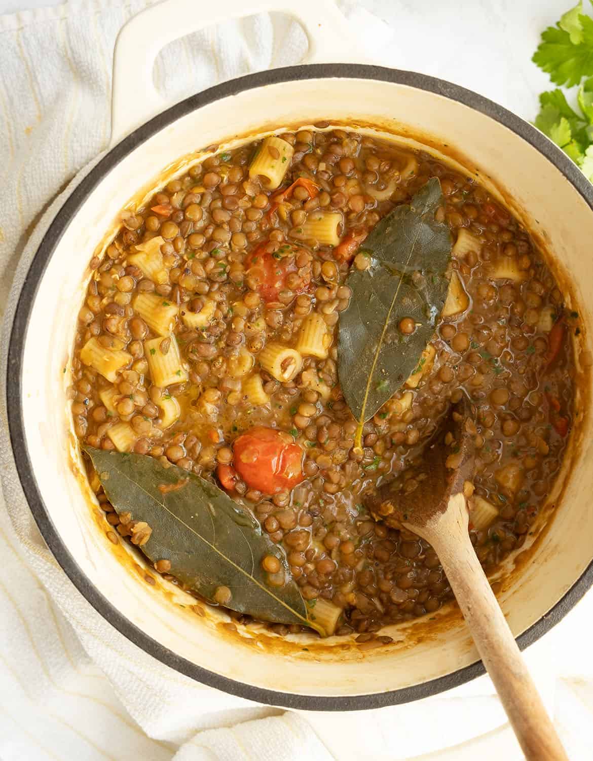 A white cast iron pot full of pasta with lentils with 2 bay leaves on top and a wooden spoon.