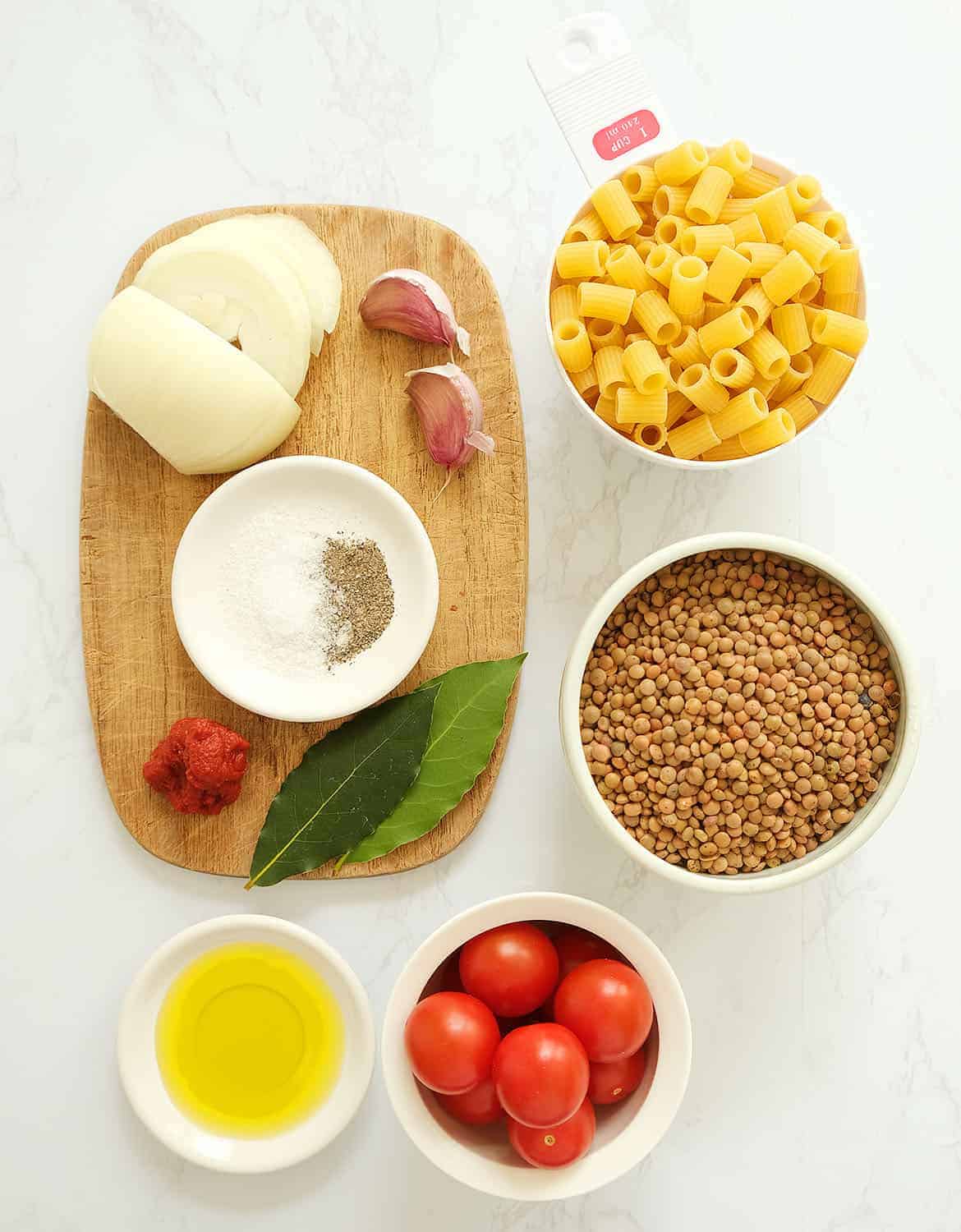 The ingredients for this pasta with lentils are arranged on a white marble table and a small wooden board.