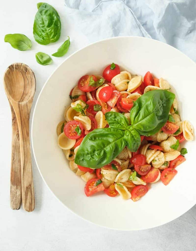 Tomato pasta salad with large basil leaves in a white salad bowl.