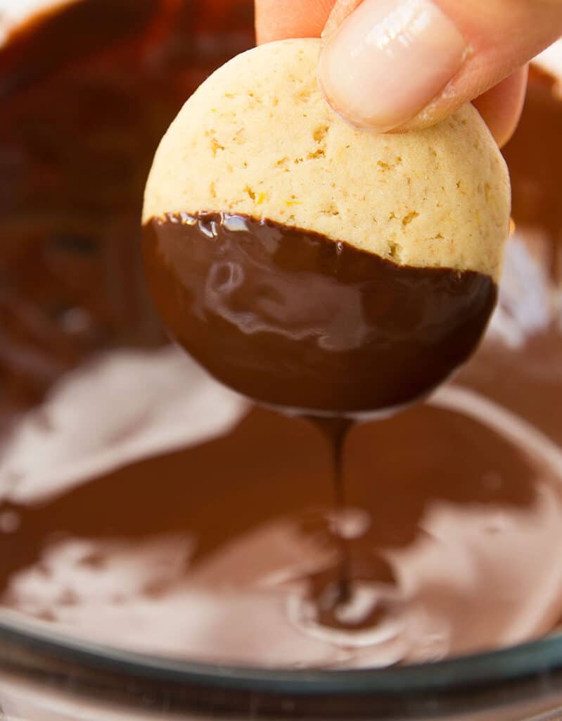 A hand dipping a round orange cookie into a bowl of melted chocolate.