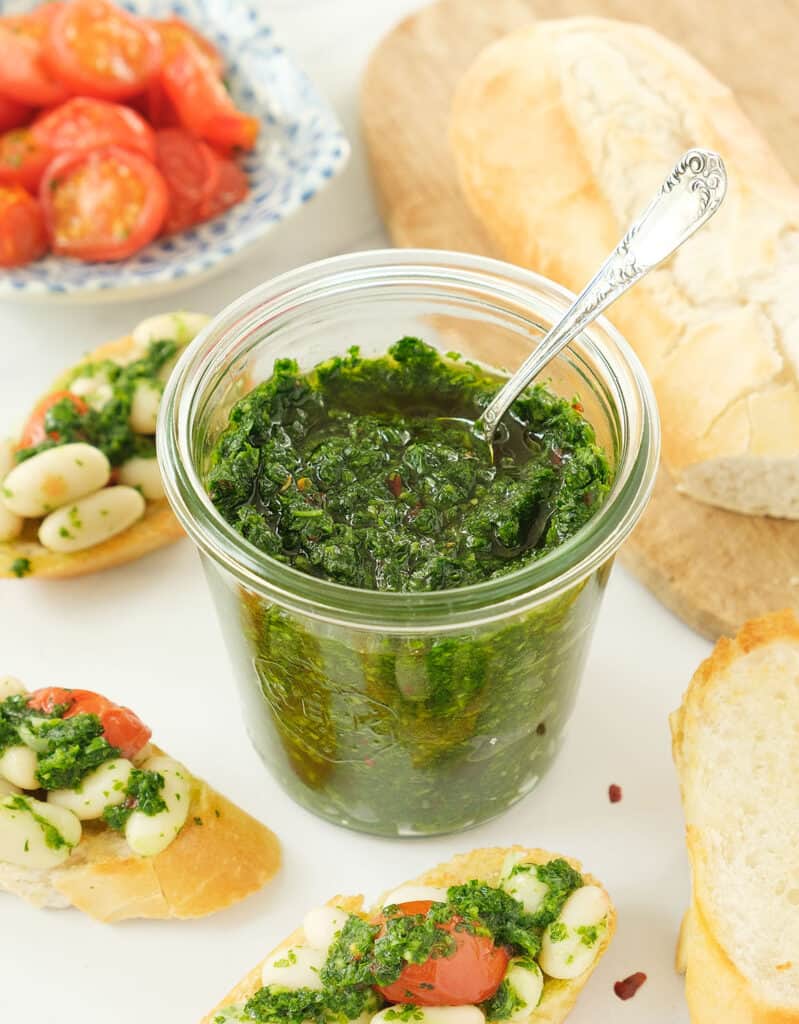A glass Mason jar full of parsley pesto; bruschetta with tomatoes, beans and pesto in the background.