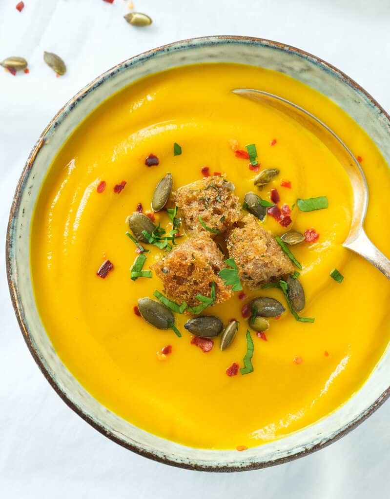 Top view of a bowl full of pumpkin carrot soup topped with croutons and garnished with pumpkin seeds and chili flakes.