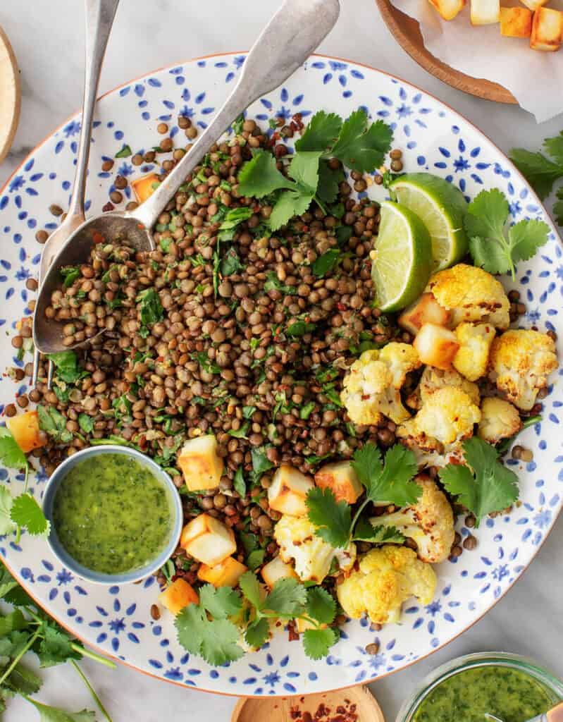 Top view of serving plate with green lentils, roasted cauliflower, coriander leaves and lime wedges.