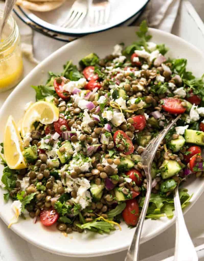 A white serving plate with green lentil salad, cherry tomatoes, crumbled feta and lemon wedges.