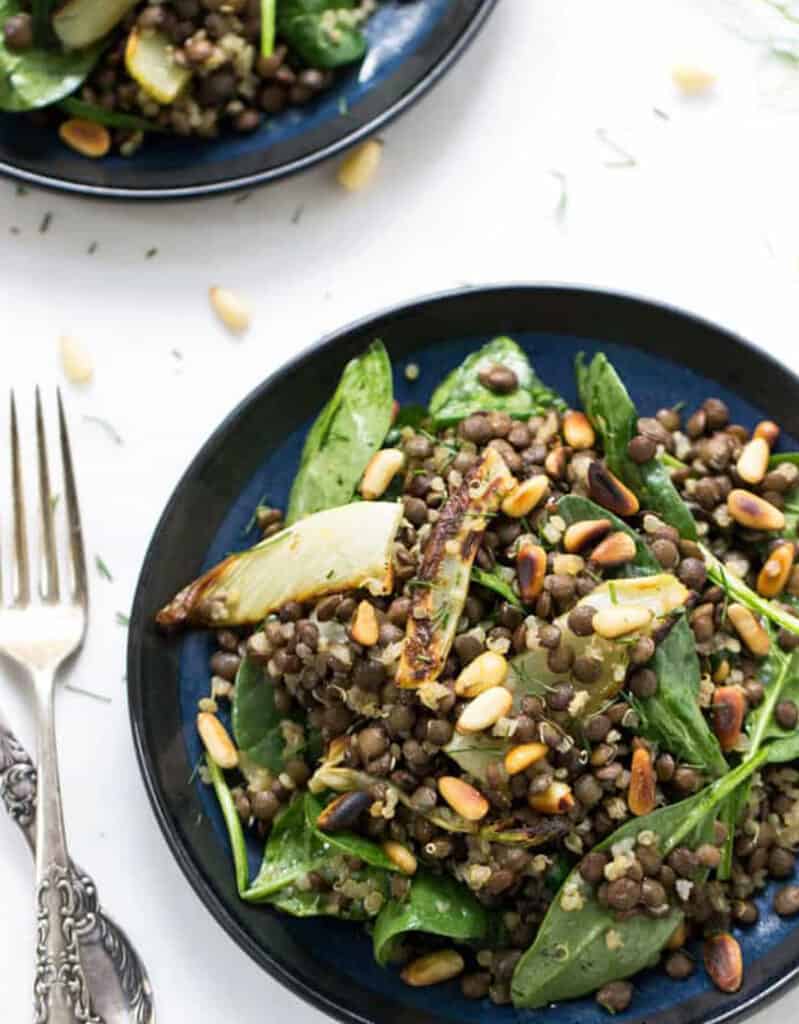 Top view of two blue plates full of green lentils salad, spinach and toasted pinenuts over a white background.