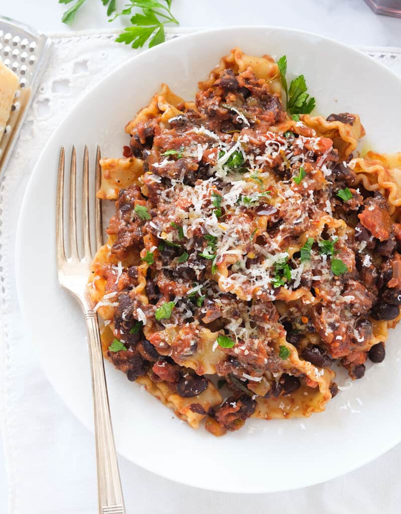 Top view of a white plate full of pasta with black beans garnished with parmesan cheese and parsley.