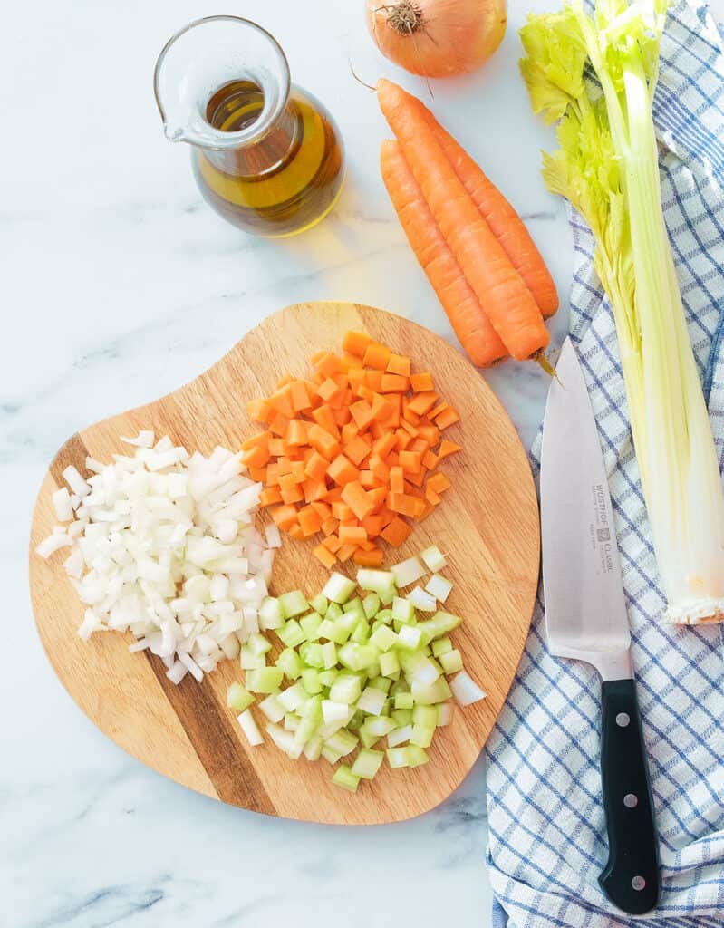 Vista dall'alto di un tagliere di legno con gli ingredienti per la ricetta del soffritto: carote tritate, cipolla, sedano e olio d'oliva.