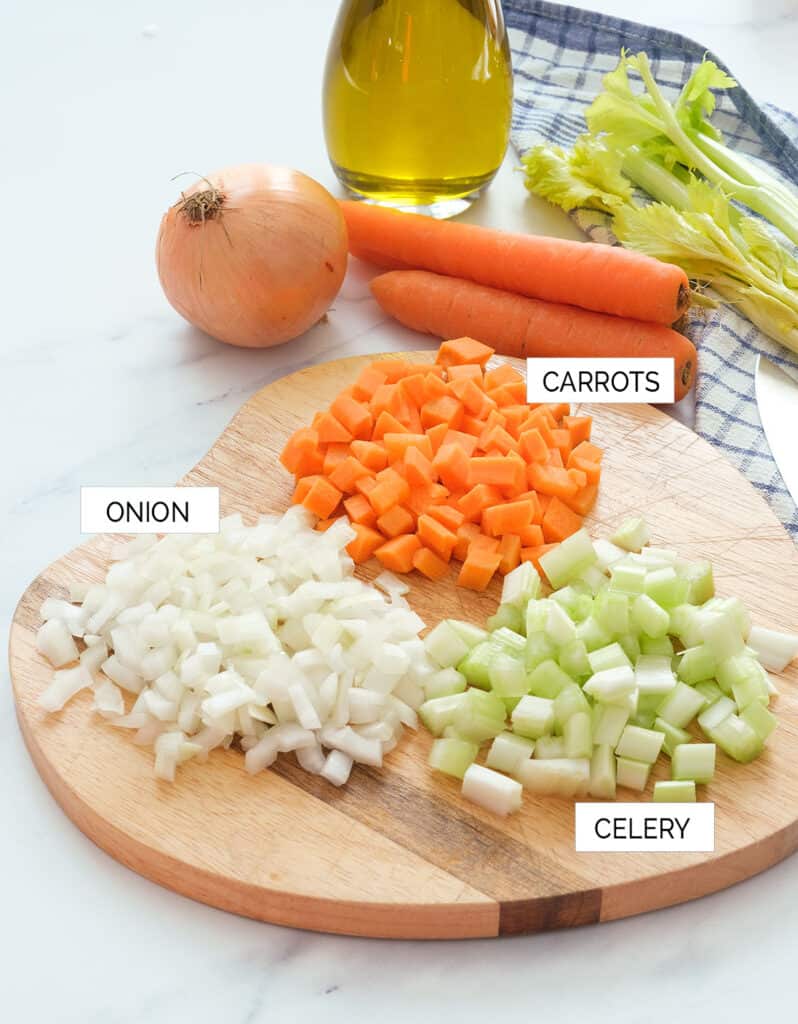 Close-up of a chopping board with the ingredients to make the Italian soffritto: onion, carrots, celery and olive oil.