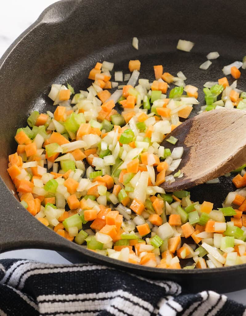 A wooden spoon stirring this soffritto recipe in a cast iron skillet. 