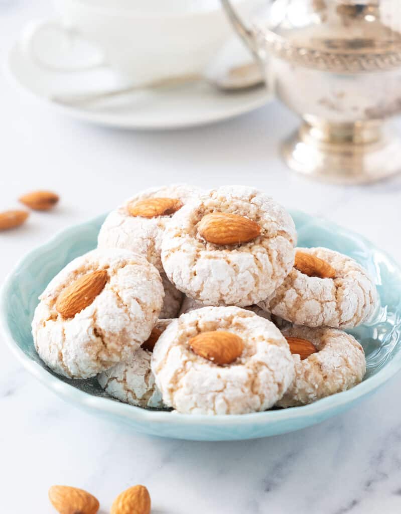 A turquoise bowl with almond cookies, and a white cup and a silver sugar bowl in the background.