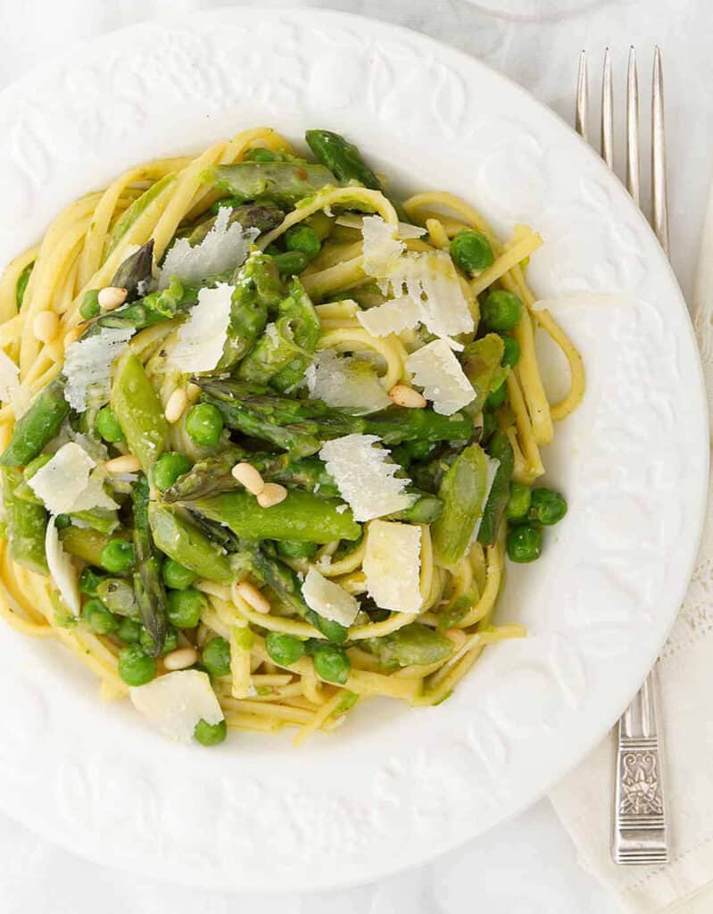 Top view of a white plate full of pasta with peas and asparagus garnished with shaved parmesan.