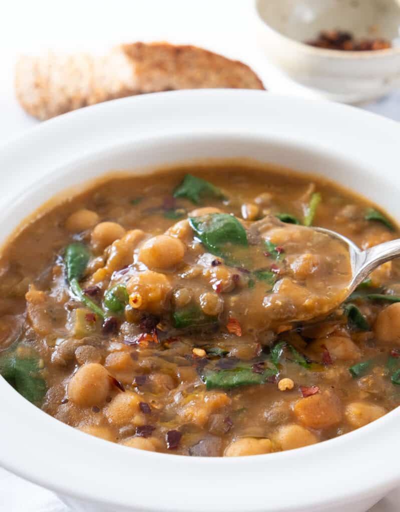 Close-up of a white bowl full of smoky chickpea and lentil soup with a spoon.