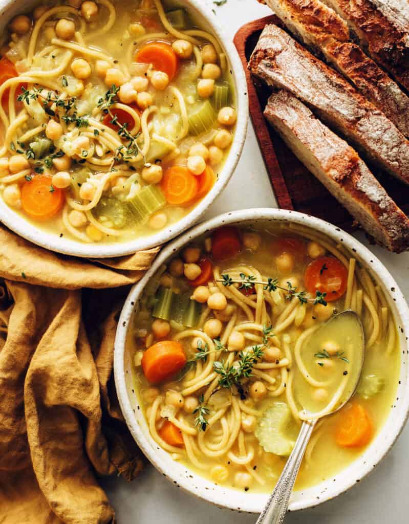 Top view of two bowls full of chickpea noodle soup with slices  of carrots and rustic bread in the background - by MInimalist Baker