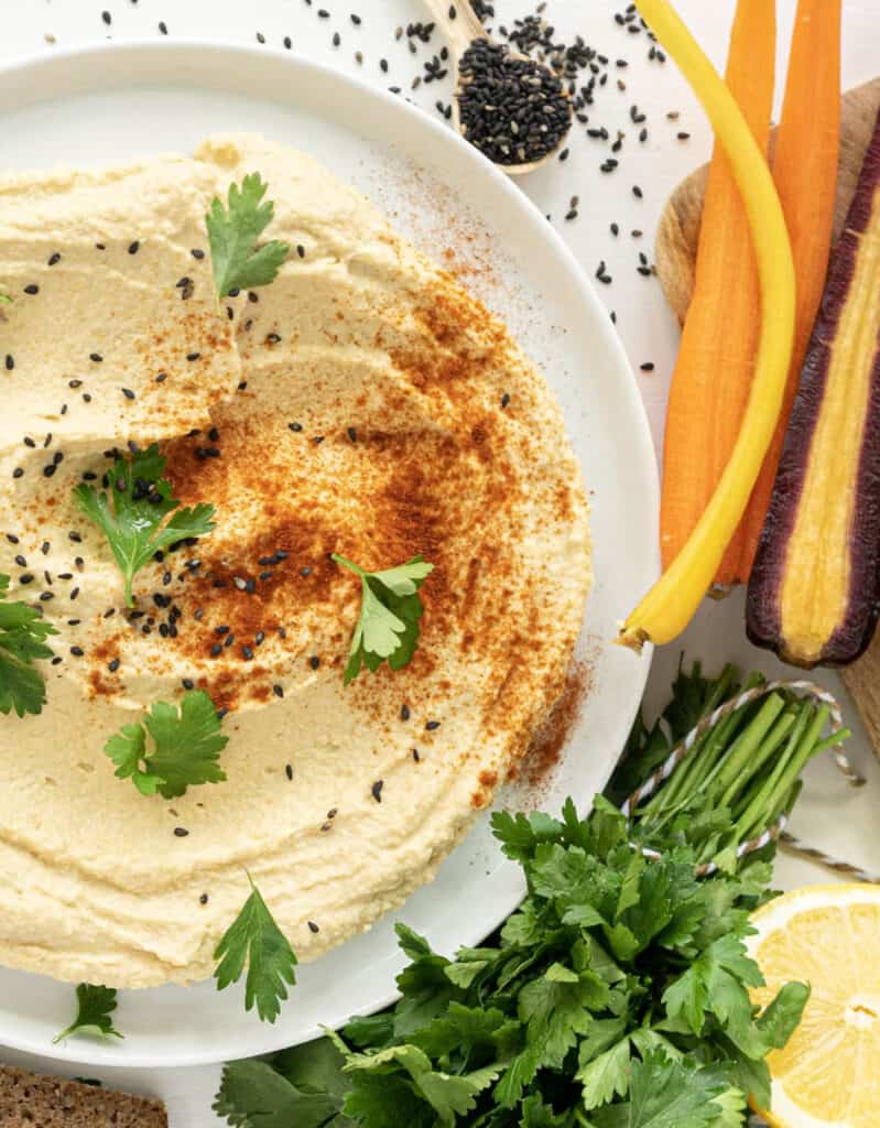 Top view of a white plate with hummus garnished with black sesame seeds and coriander. Slices of carrots next to the plate - by Gathering Dreams