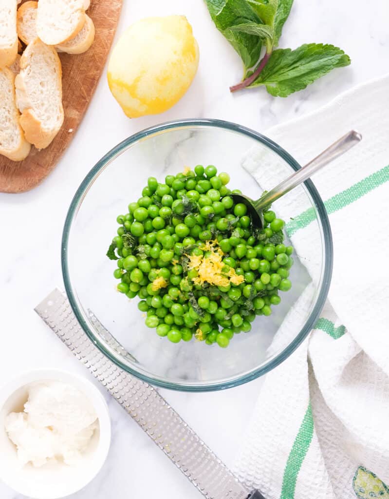 Top view of a glass bowl full of peas near crostini and a small bowl full of ricotta.