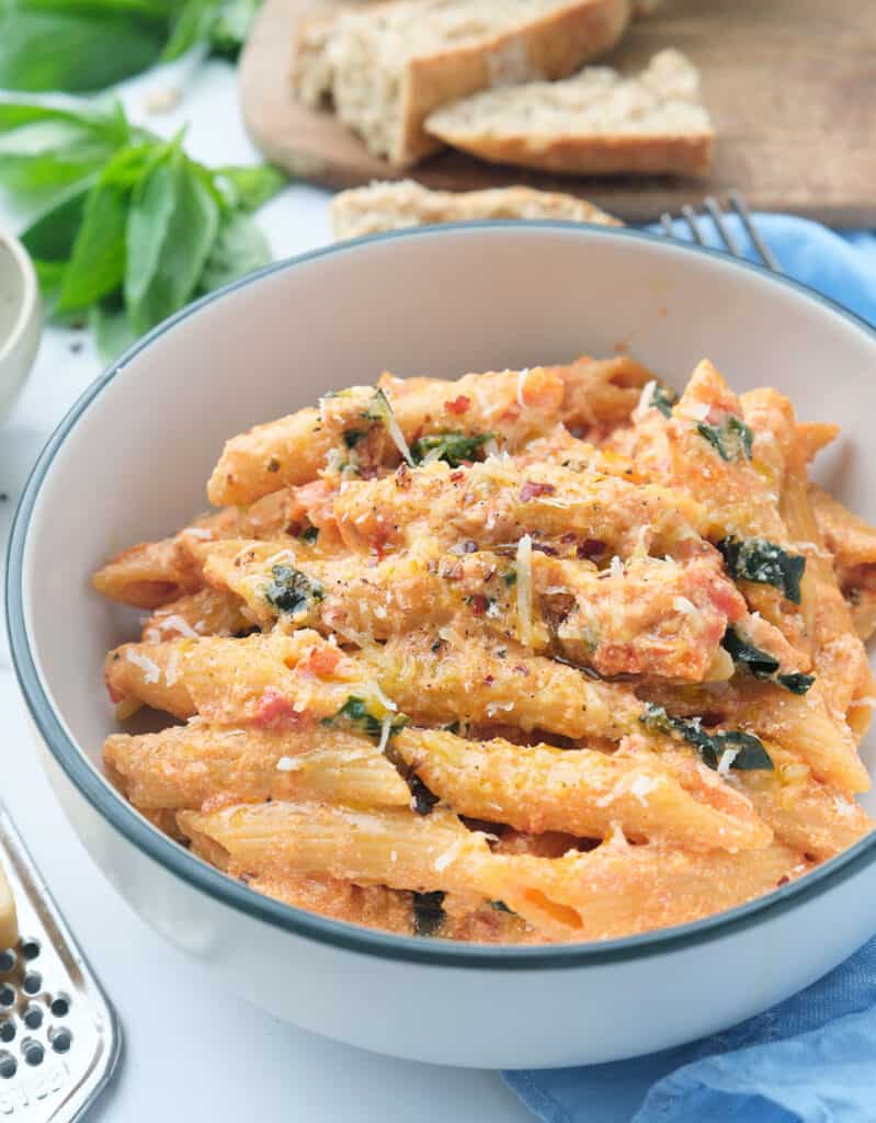 A bowl full of penne pasta with basil tomato sauce, basil leaves and slices of bread in the background.