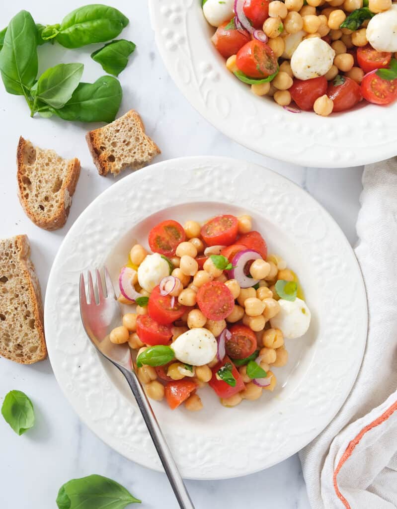Top view of two white plate full of Italian chickpea salad with tomatoes and mozzarella, slices of bread and basil leaves in the background.